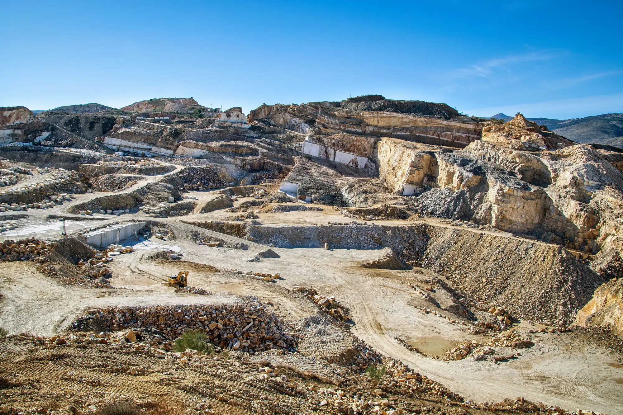 A working marble quarry seen from above its rim, cut blocks stacked along haul roads and one excavator on the floor